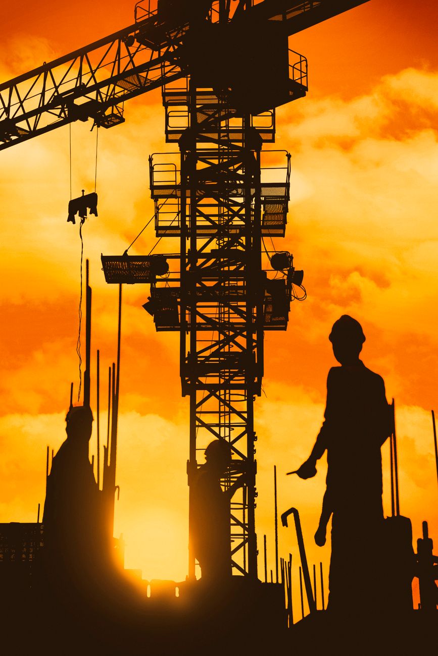 Construction workers and a crane silhouetted against a sunset on an industrial site